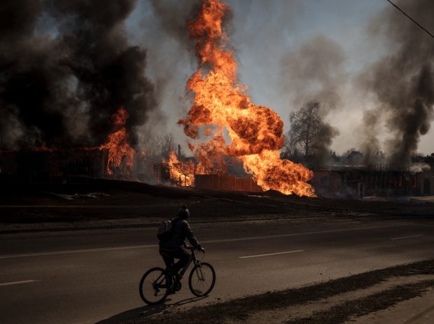 Um homem passa de bicicleta por lojas e casas em chamas após um ataque russo em Kharkiv. Foto de Felipe Dana/AP - 2022